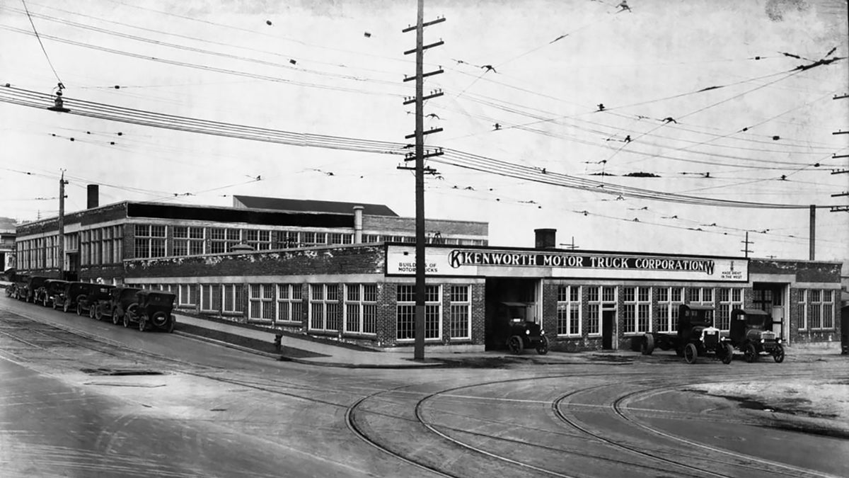 Black and white photo of the Kenworth Motor Truck Corporation building on a street corner, with several early 20th-century trucks parked outside and overhead utility wires crossing above.