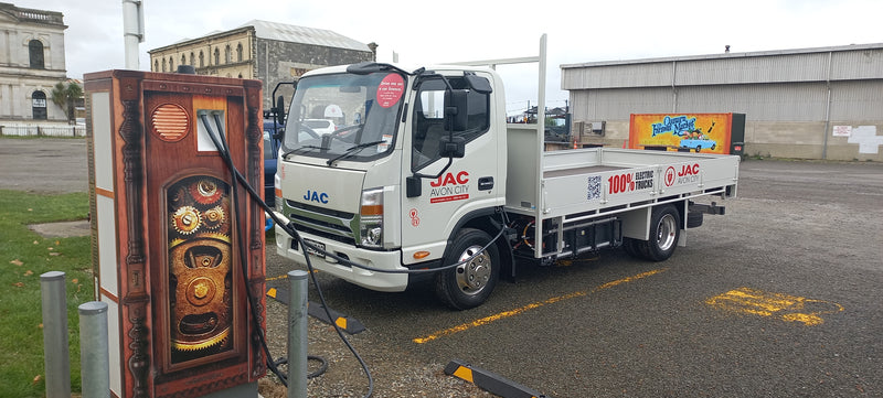 A white JAC electric truck is parked and plugged into a charging station decorated with a steampunk design. The truck is in a parking lot near industrial buildings, with a mural visible in the background.