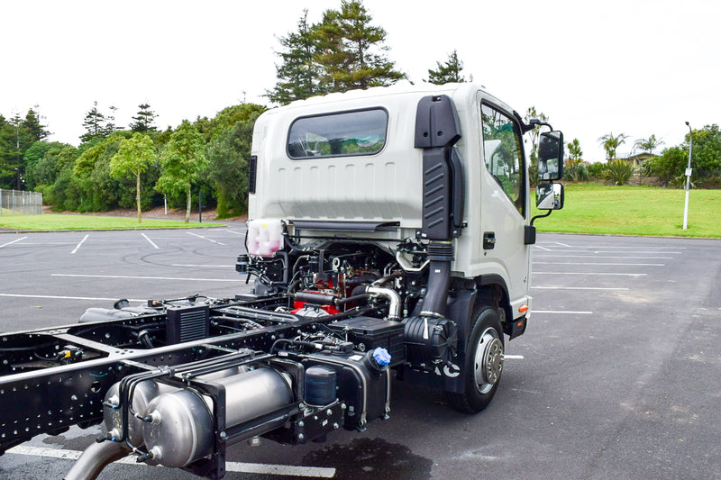 A white commercial truck with its cab tilted forward, exposing the engine and chassis components, is parked in an empty lot surrounded by greenery and trees.