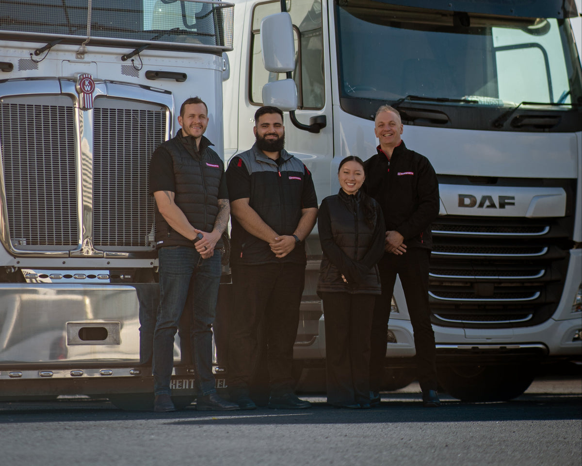 Four people in matching black vests stand smiling in front of two large white trucks parked side by side on a sunny day.