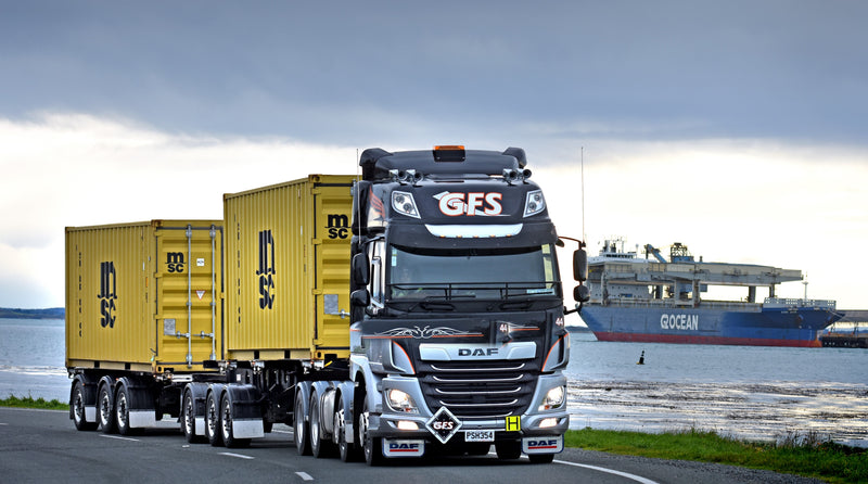A large DAF truck with GES branding hauls two yellow MSC shipping containers along a road near a body of water, with a cargo ship and port facilities visible in the background under an overcast sky.