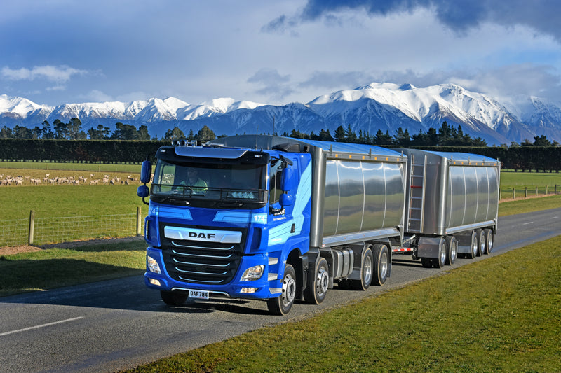 A blue DAF truck with a large silver trailer drives on a rural road. Snow-capped mountains and a green field with sheep are visible in the background under a partly cloudy sky.
