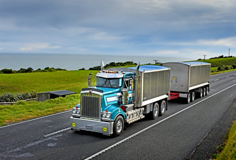 A shiny blue semi-truck with two silver trailers drives along a rural highway bordered by green fields, with the ocean and a cloudy sky visible in the background.