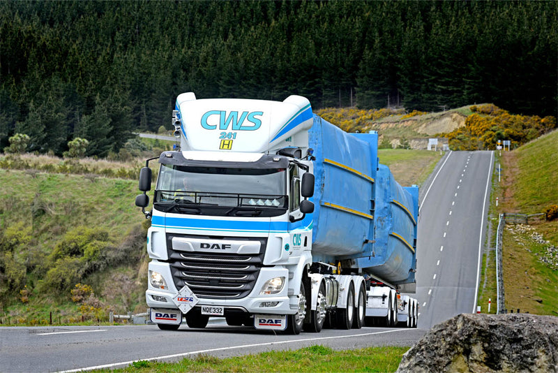 A large white and blue DAF truck with CWS 24/7 on the cab hauls two oversized, blue-wrapped loads along a country road flanked by grassy hills and dense green forest.