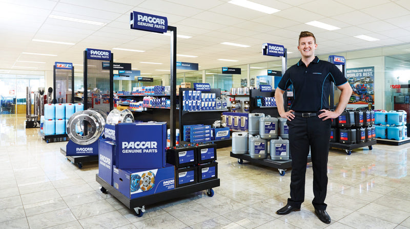 A smiling man in a black uniform stands in a well-lit auto parts store with shelves displaying PACCAR Genuine Parts, oil containers, and automotive products. The store has shiny tile floors and modern, organized displays.