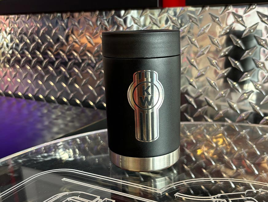 The STUBBY KW CAN COOLER, a black insulated tumbler with a silver KW logo, sits on a reflective surface before a metal diamond plate backdrop—perfect for keeping your drinks cold for hours.