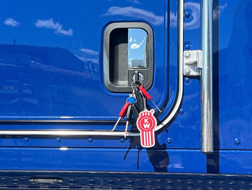 A KEYRING KW RUBBER with red Kenworth Bug logo hangs from the door handle of a blue truck, its shiny surface reflecting clouds.
