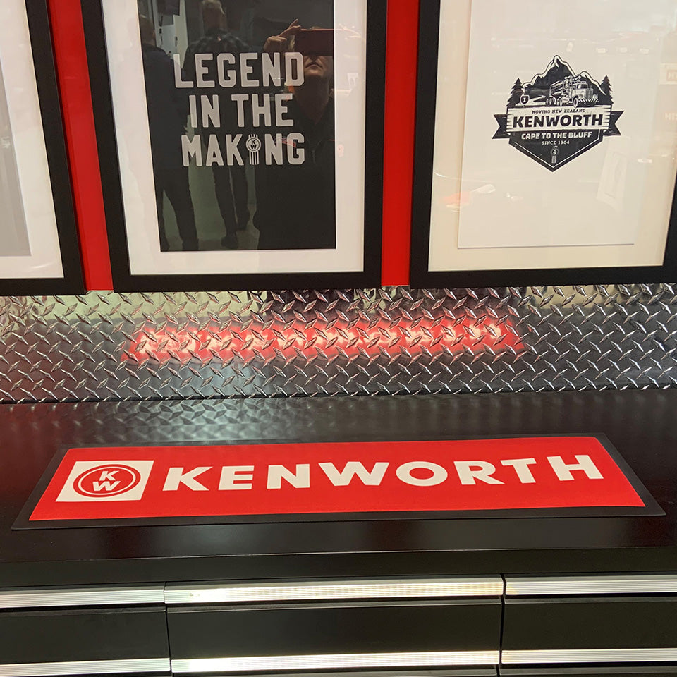 A framed Kenworth poster and a truck graphic decorate the metal wall. On the counter, the BAR MAT RED KENWORTH brings style and function to your home bar setup, complemented by a bold red Kenworth sign.