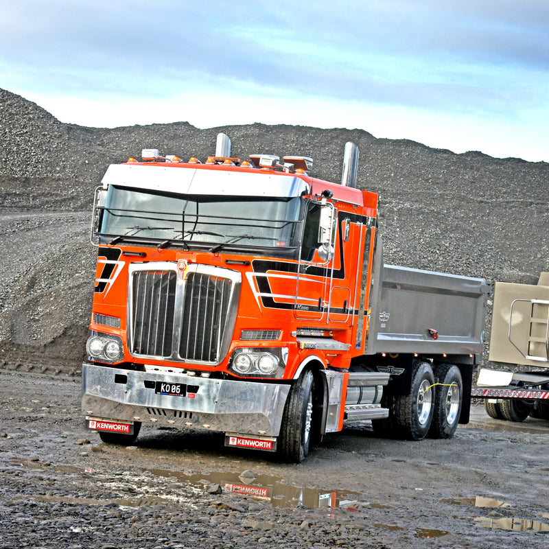 A bright orange Kenworth dump truck with chrome details is parked on a muddy construction site, with gravel piles and another truck visible in the background.