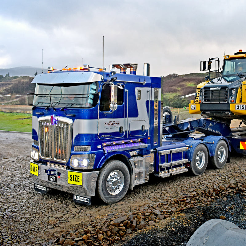 A large blue and silver semi-truck labeled OVERSIZE is parked on a gravel area, carrying heavy construction equipment on a trailer in a rural, hilly landscape.