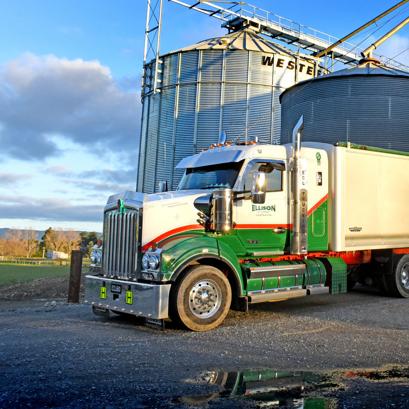 A green and white semi-truck with a trailer is parked on gravel near large metal grain silos at a farm. The scene is lit by low evening sunlight, with blue sky and clouds in the background.