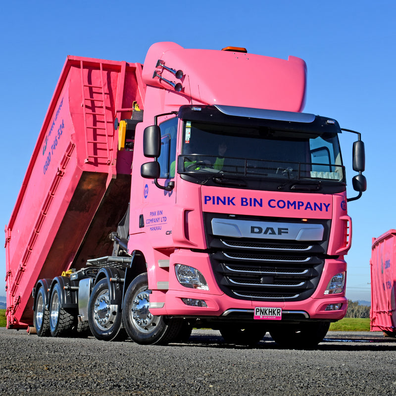 A large pink DAF truck with PINK BIN COMPANY on the front is unloading a pink dumpster container. The scene is outdoors on a bright, clear day. The truck has a visible license plate reading PNKHKR.