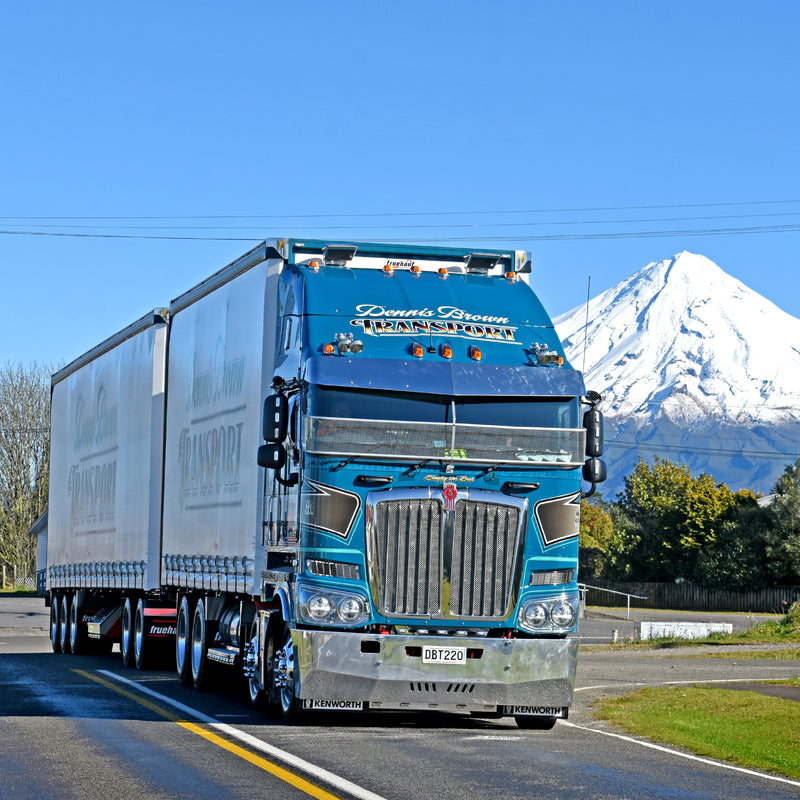 A large blue semi-truck with a trailer drives on a road with snow-capped Mount Taranaki and green trees visible in the background on a clear, sunny day.