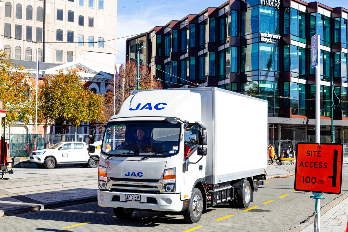 A white JAC delivery truck drives down a city street with modern glass buildings in the background and a red Site Access 100m sign visible on the right side.