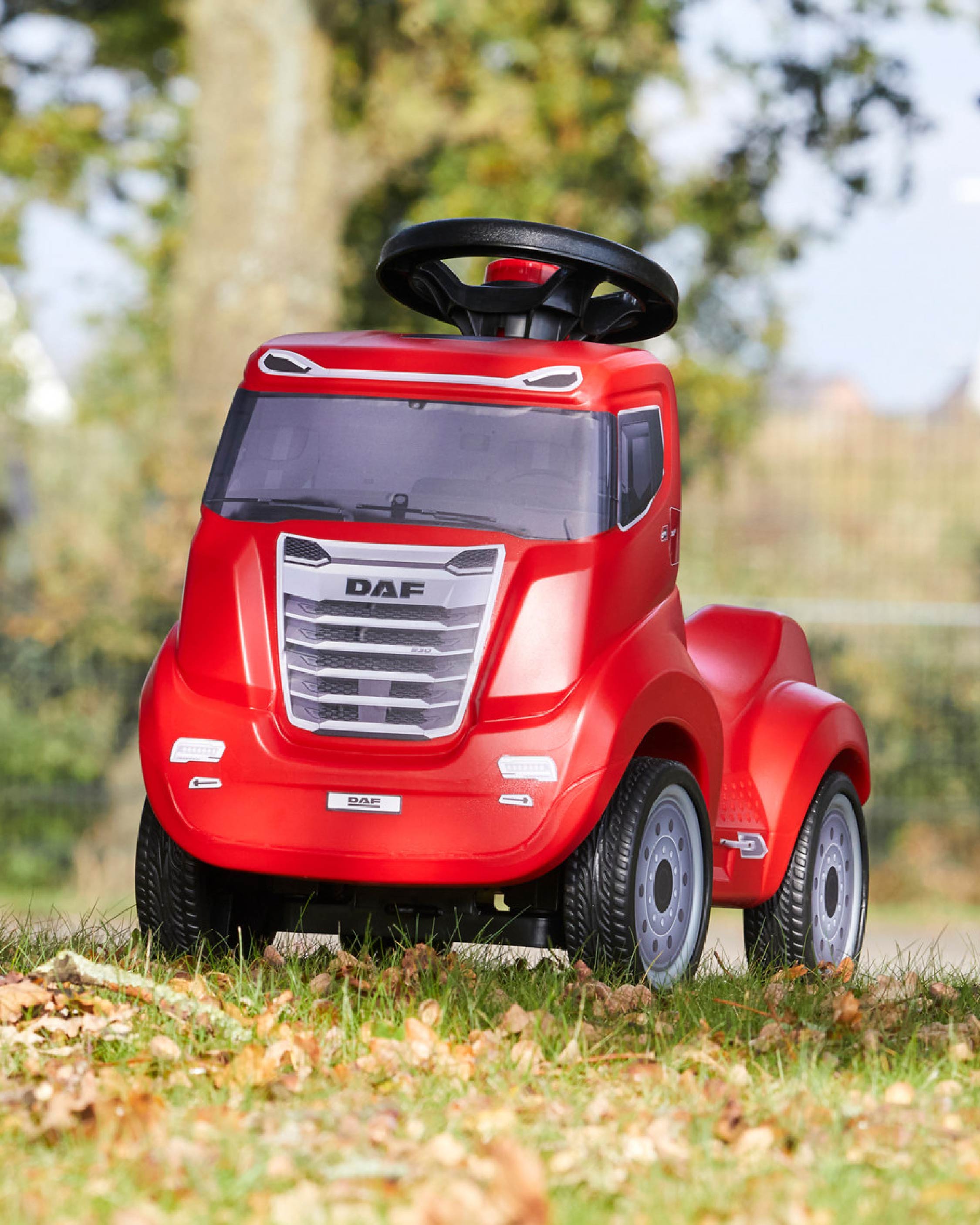 A small, red toy truck with a large steering wheel sits on grass and fallen leaves outdoors, with trees and a blurred fence in the background.