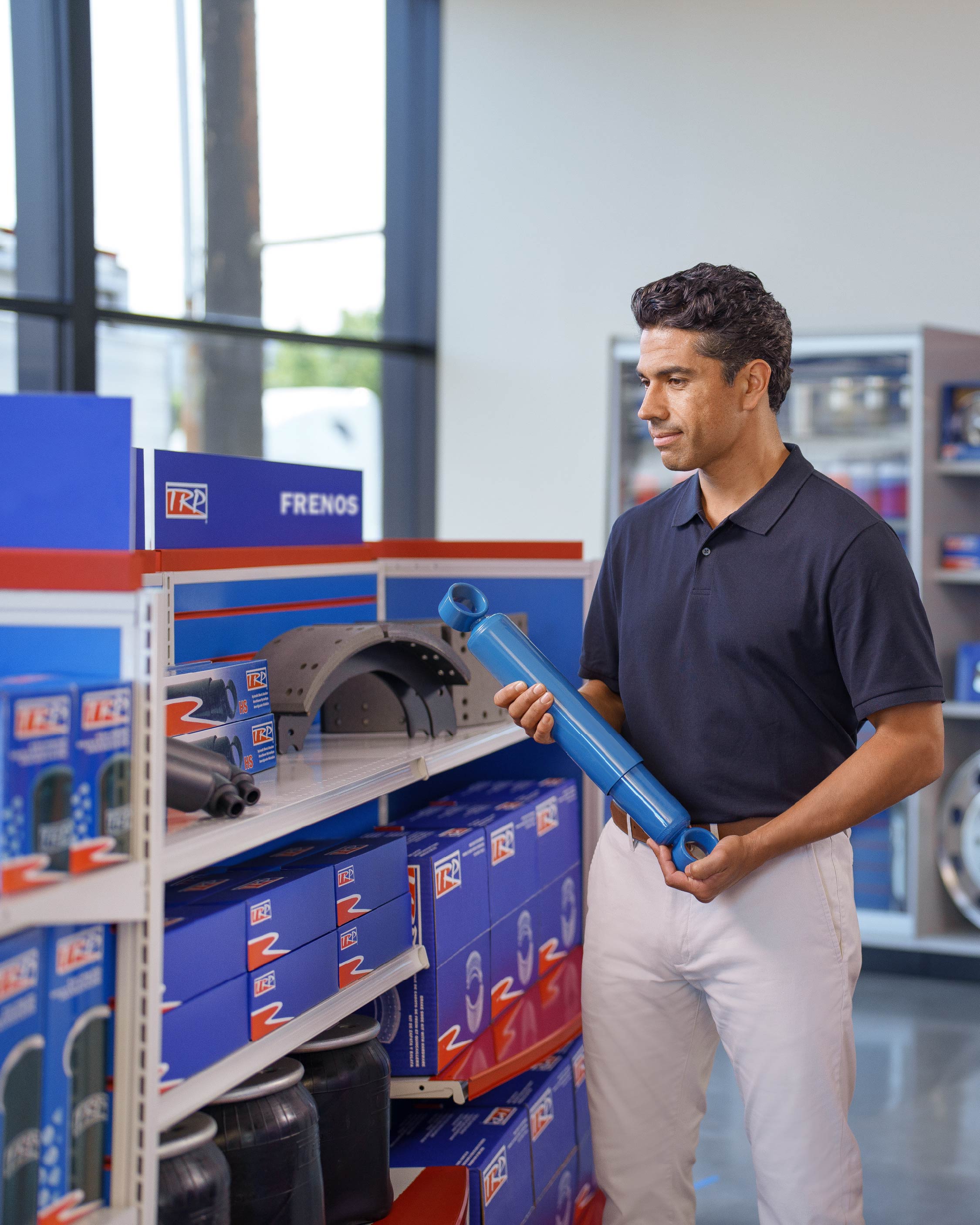 A man in a navy polo and white pants inspects a blue automotive part in an auto parts store aisle labeled Frenos, surrounded by shelves stocked with various car brake components.
