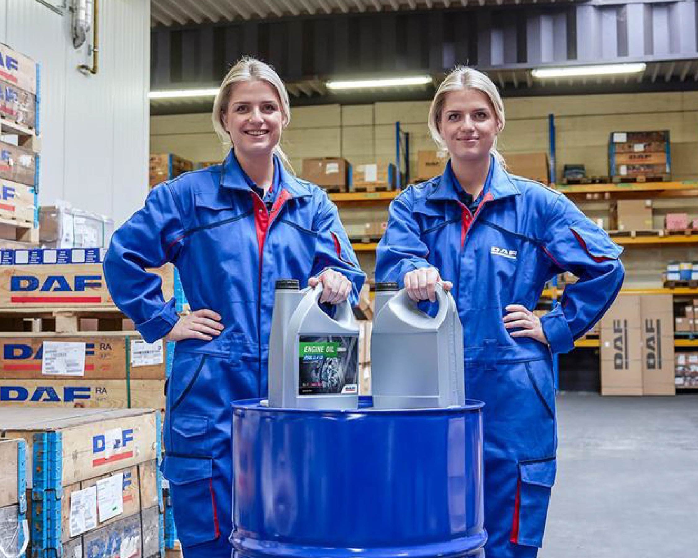 Two people in blue coveralls stand in a warehouse, each resting a hand on large oil containers placed on top of a blue barrel. Stacks of boxes and shelves filled with items are visible in the background.