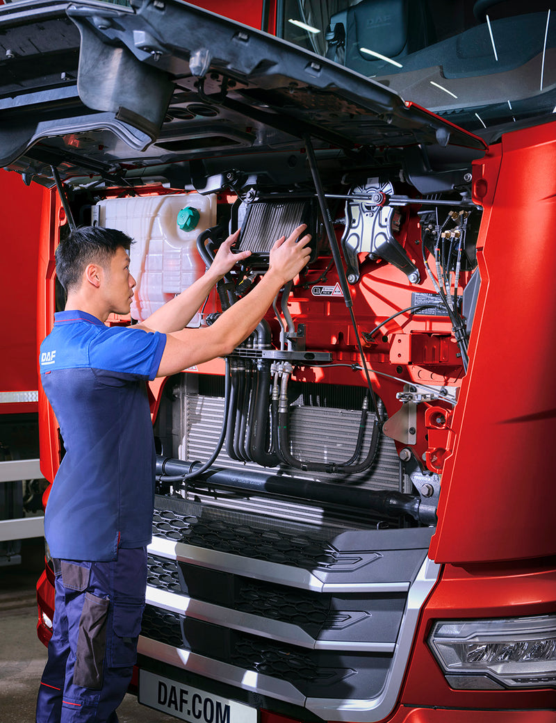 A mechanic in a blue uniform inspects or repairs the engine of a red DAF truck with its hood open, focusing on components inside the engine compartment.
