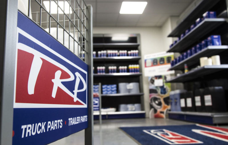Interior of a store with shelves stocked with truck and trailer parts. A sign in the foreground reads TRP TRUCK PARTS TRAILER PARTS. The floor has branded mats, and the room is well-lit with overhead lights.