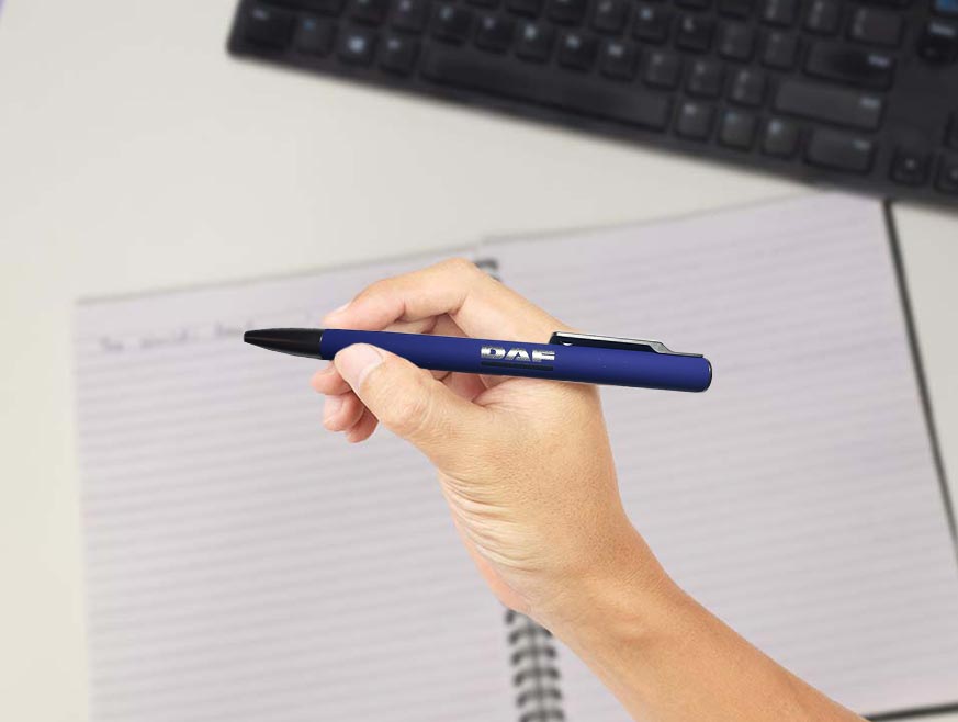A hand holds the eco-friendly PEN DAF BLUE above an open lined notebook with faint writing, while a black computer keyboard can be seen in the background.