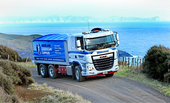 A large blue and white Auckland Landscape Supplies truck drives along a gravel road with coastal hills and the ocean in the background.