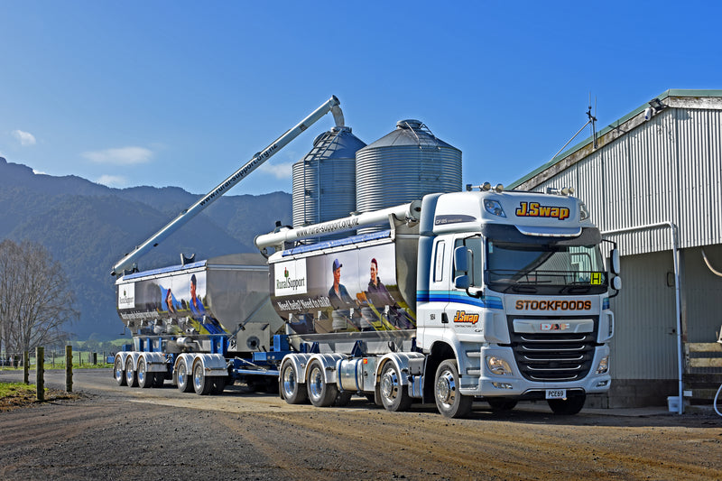 A large J.Swap Stockfoods truck with two trailers is positioned beside a building, unloading feed into metal silos via a long overhead pipe on a rural property with mountains in the background.