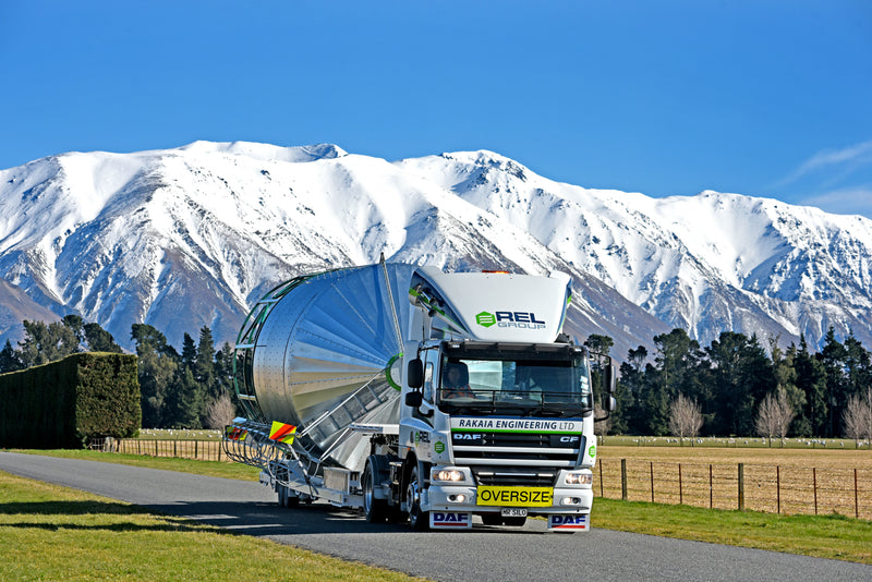 A large white truck labeled OVERSIZE transports a massive metal structure on a rural road, with snowy mountains and green fields visible under a clear blue sky in the background.