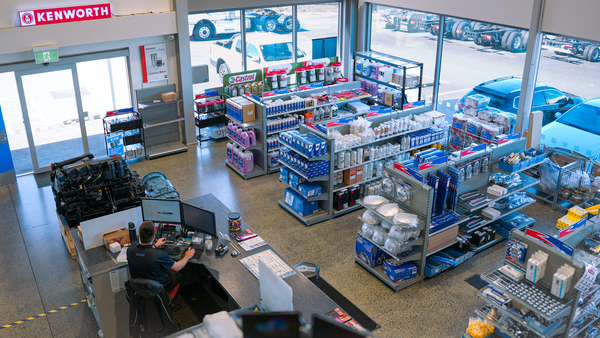 An overhead view of a truck parts store shows shelves stocked with auto parts and oils, a counter with a seated employee on a computer, and large windows displaying vehicles parked outside.