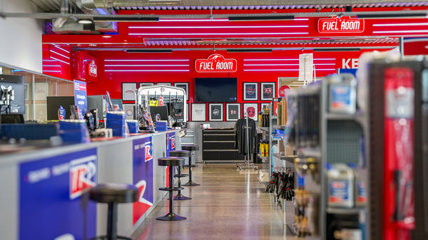 A brightly lit auto parts store interior with a red Fuel Room sign above the counter, stools, shelves stocked with products, and framed items displayed on the back wall.