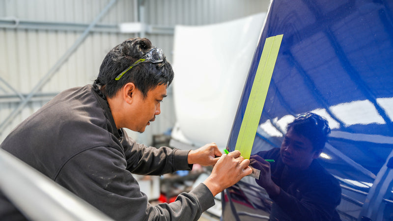 A person wearing safety glasses is carefully applying green masking tape to the edge of a glossy blue car panel in an automotive workshop.