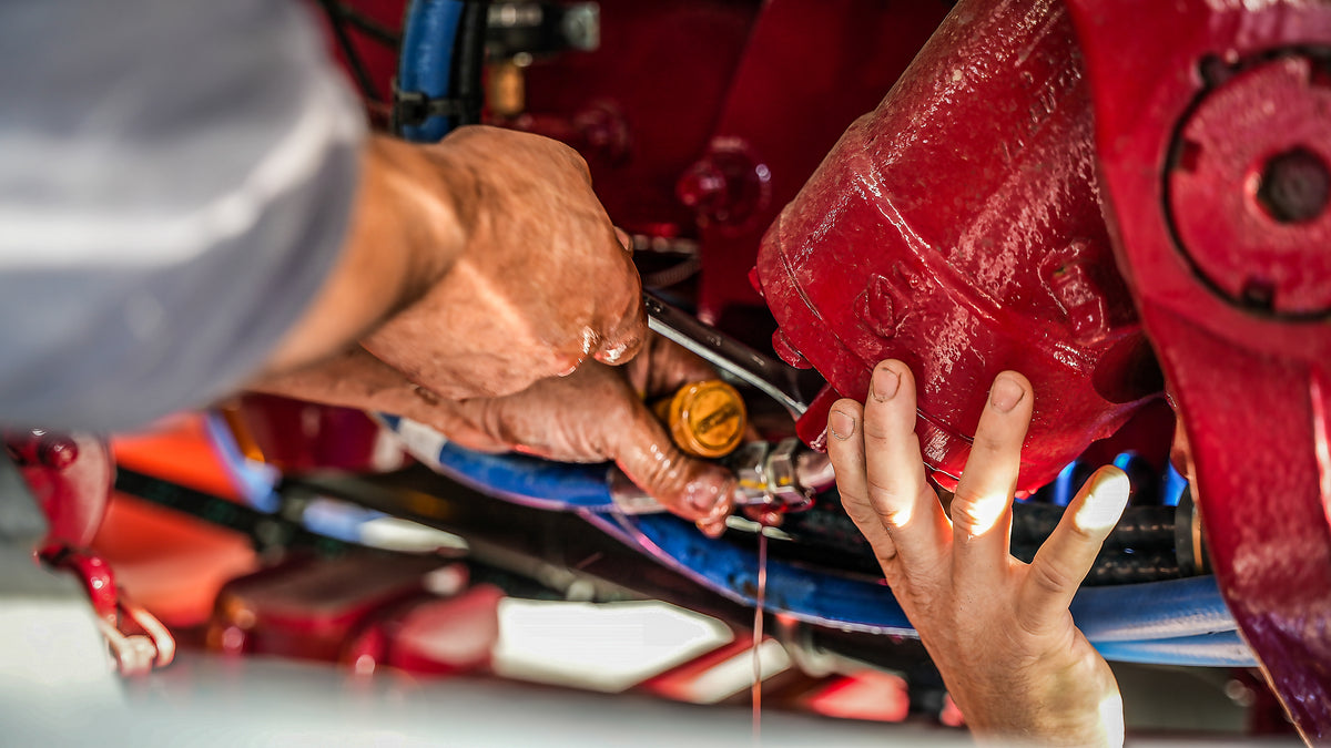 Close-up of two people’s hands working on a red engine, using tools to adjust or repair a component while oil or fluid drips down from the machinery. Blue hoses and engine parts are visible in the background.