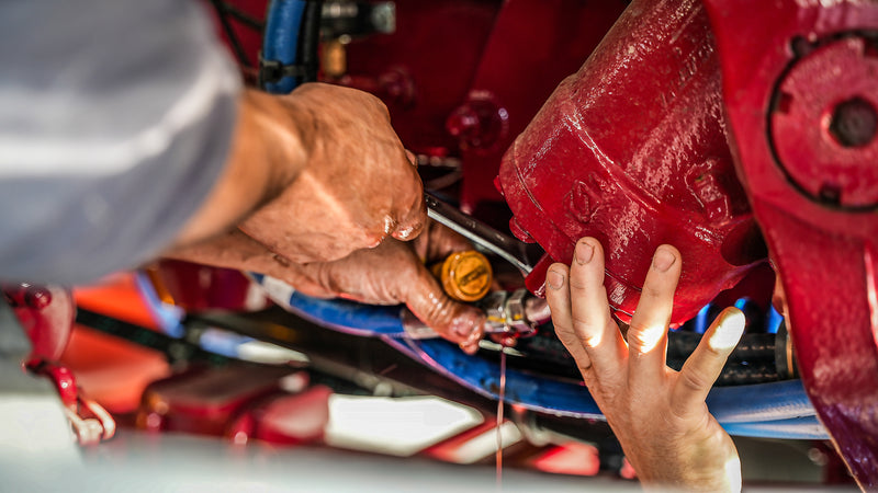 Close-up of two people’s hands working on a red engine, using tools to adjust or repair a component while oil or fluid drips down from the machinery. Blue hoses and engine parts are visible in the background.