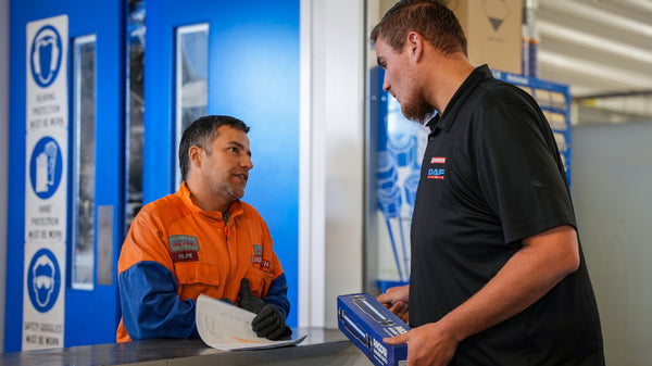 A man in an orange safety suit speaks with another man in a black shirt holding a box across a counter in an industrial setting with blue doors and safety signs in the background.