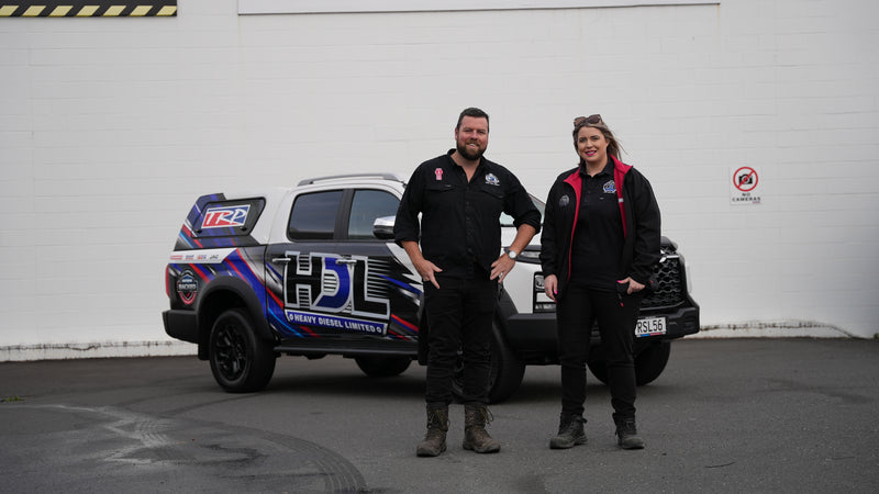 Two people stand smiling in front of a pickup truck with colorful decals and HDL Sheaf Diesel Limited branding, parked on a paved surface with a white building and no parking sign in the background.