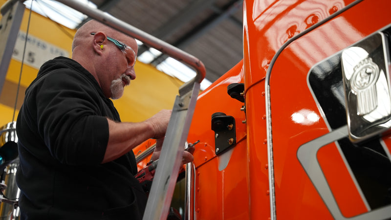 A man wearing safety glasses and ear protection works on the side of a shiny orange truck, using tools while standing on a step ladder inside an industrial building.