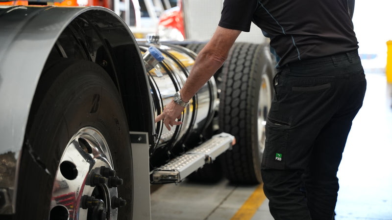 A person wearing black pants and a black shirt points at a shiny metal tank attached to a large commercial truck, which is parked indoors on a concrete floor with yellow safety lines.