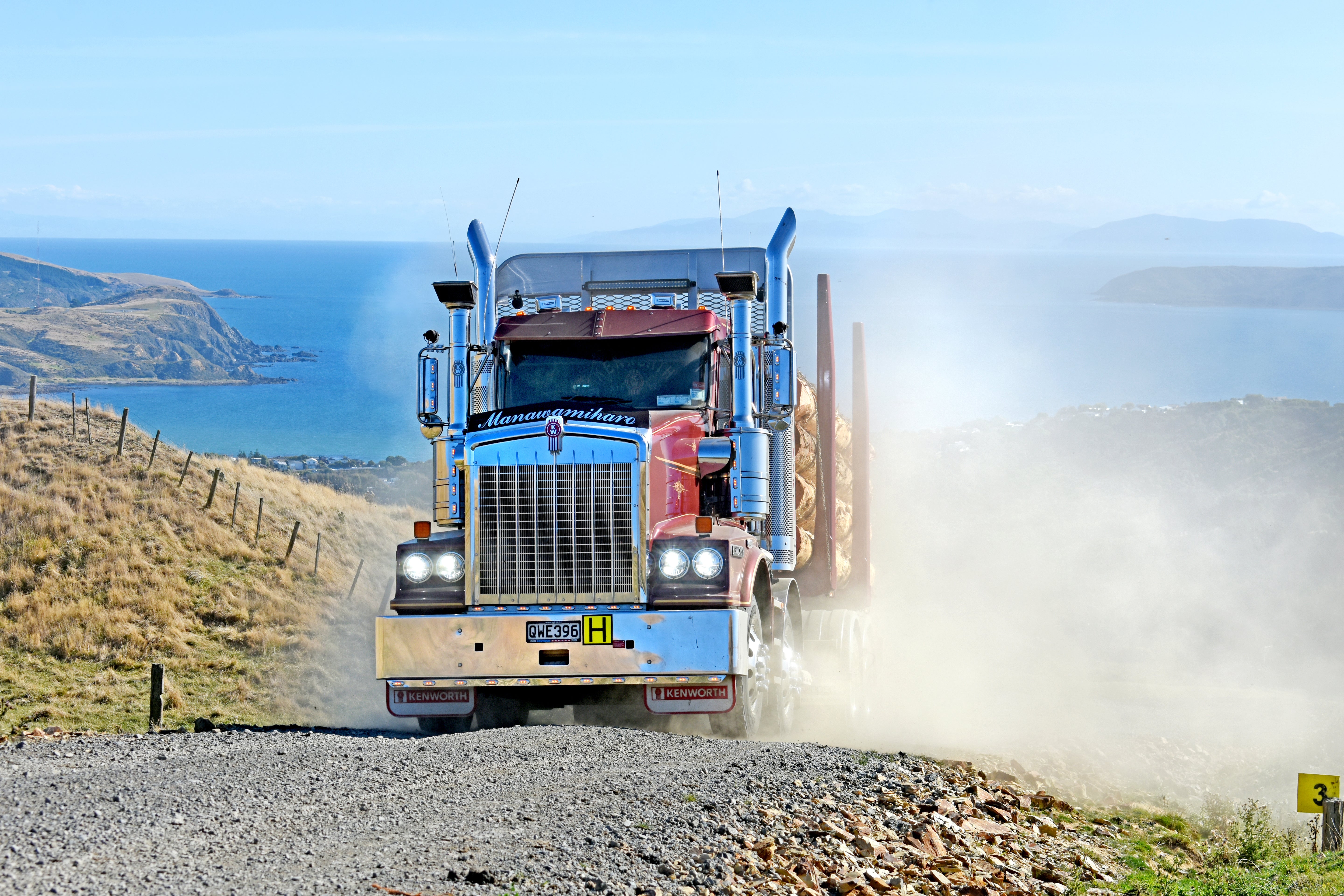 A large semi-truck hauling logs drives on a gravel road, kicking up dust, with grassy hills and a blue ocean coastline in the background under a clear sky.