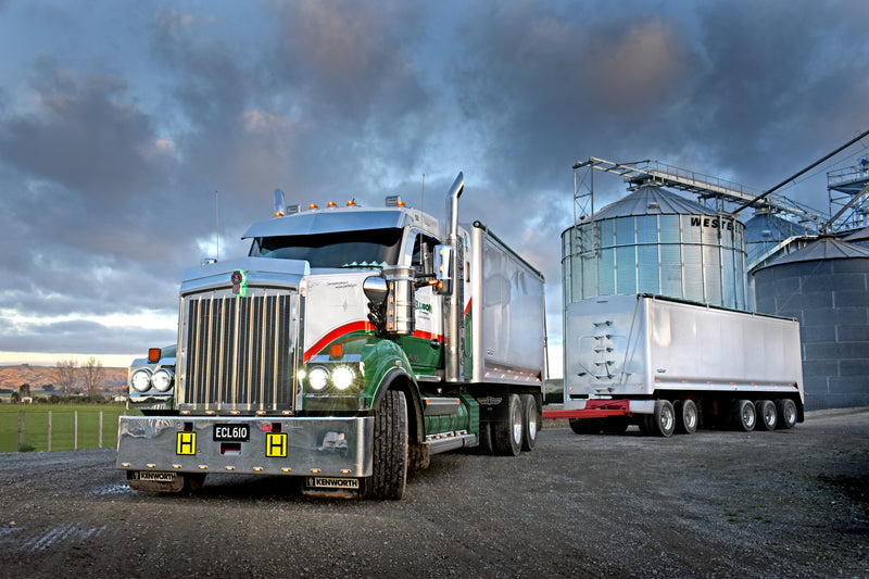 A large green and white semi-truck with a double trailer is parked on a gravel road near tall metal silos under a cloudy sky. The trucks cab door is open, and the headlights are on.