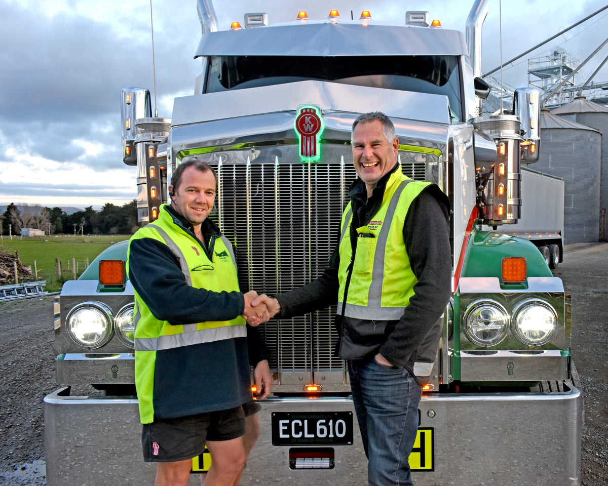 Two men wearing high-visibility vests shake hands and smile while standing in front of a large green and silver semi-truck. One man is in shorts; the other is in jeans. The scene is outdoors, with cloudy skies.