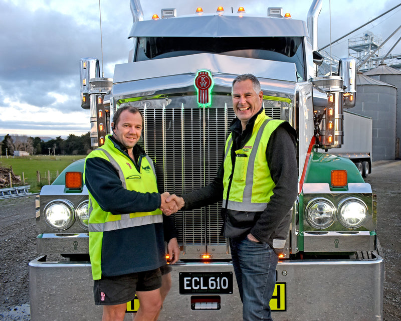 Two men wearing high-visibility vests shake hands and smile while standing in front of a large green and silver semi-truck. One man is in shorts; the other is in jeans. The scene is outdoors, with cloudy skies.