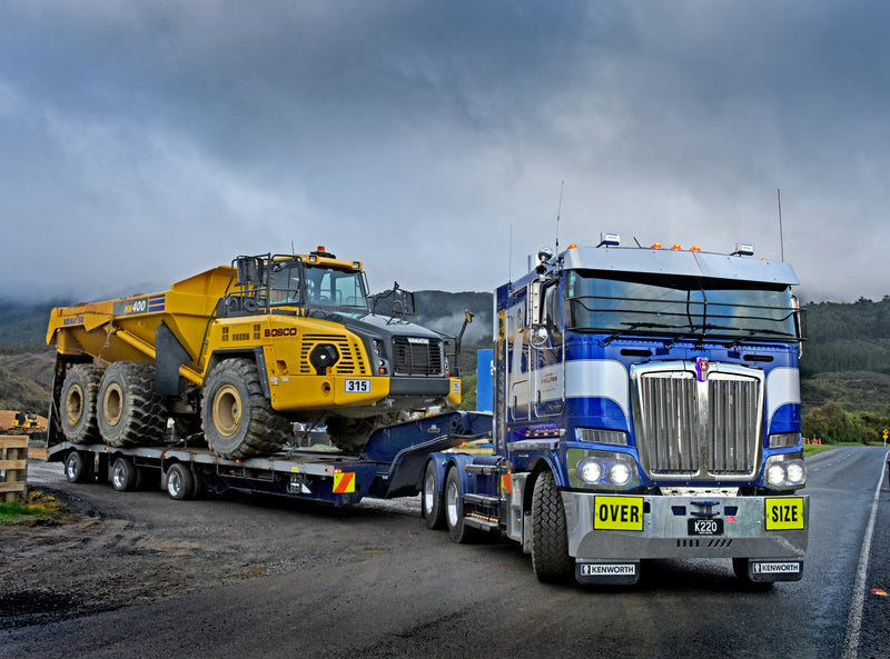 A large blue and white truck labeled OVER SIZE transports a yellow articulated dump truck on a flatbed trailer along a rural road, with cloudy skies and hills in the background.