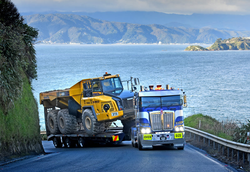 A large blue and white truck transports a yellow dump truck on a trailer along a coastal road, with the ocean and distant hills in the background. The OVER SIZE sign is visible on the trucks front.