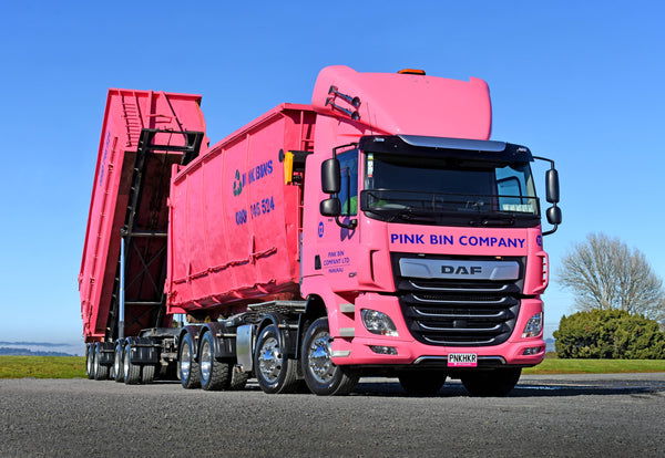 A large, bright pink DAF truck labeled PINK BIN COMPANY with a matching pink bin trailer is parked on a paved surface under a clear blue sky; the trailer is raised at an angle.