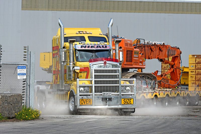 A large yellow Kenworth truck labeled OVER SIZE hauls heavy equipment, raising dust as it drives through an industrial area with a metal fence and warehouse in the background.