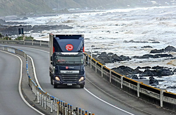 A large truck drives along a coastal road bordered by rocky shoreline and rough waves, with cloudy skies and hills in the background.