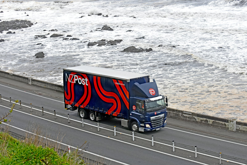 A blue NZ Post delivery truck with red and white accents drives along a coastal road next to the ocean, with waves crashing against rocks in the background.
