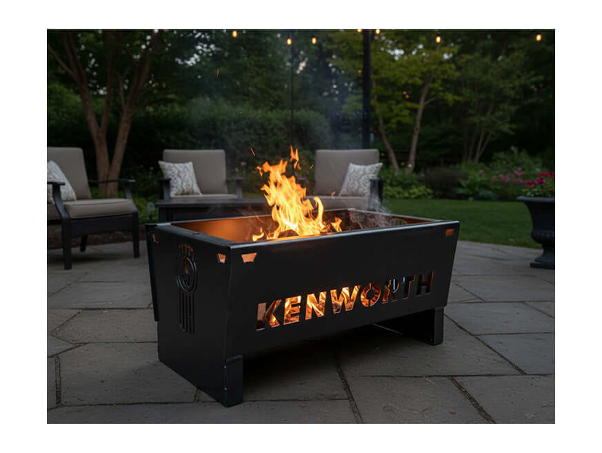 The FIREPIT KW STEEL, a black rectangular steel firepit with "KENWORTH" cut out, glows warmly in a backyard. Patio chairs and greenery surround it under twinkling string lights for a cozy outdoor vibe.