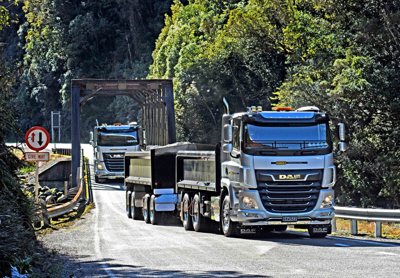 Two large DAF trucks with trailers drive on a winding rural road, approaching a narrow steel bridge surrounded by trees. A Give Way traffic sign is visible on the left side of the road.