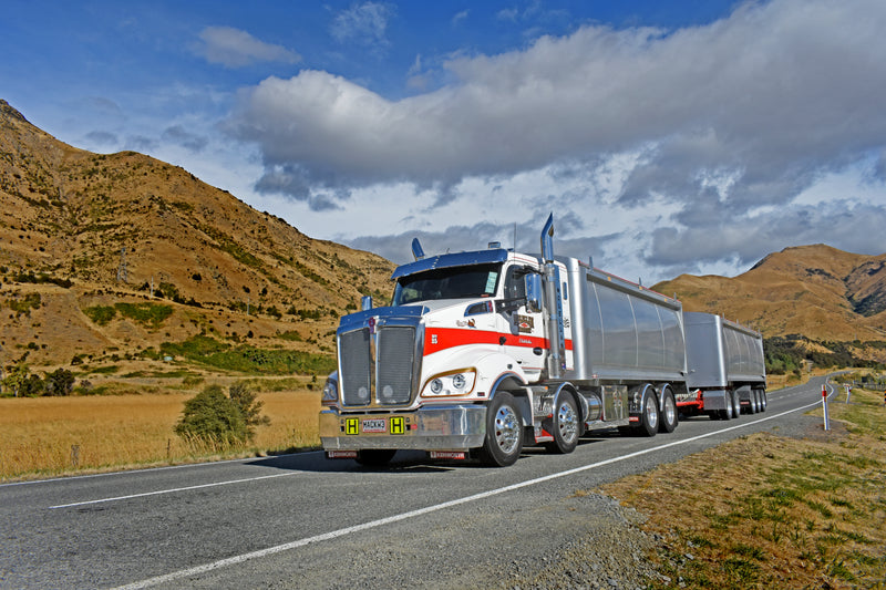 A large silver and white semi-truck with a trailer drives on a rural road, with dry grassy hills and partly cloudy skies in the background.