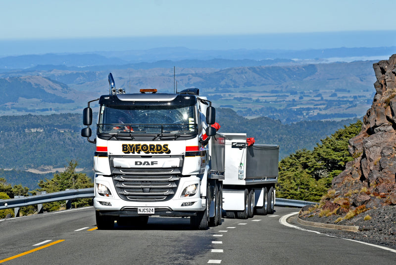 A large white BYFORDS DAF truck with a trailer drives uphill on a winding road, surrounded by greenery and distant mountains under a blue sky.